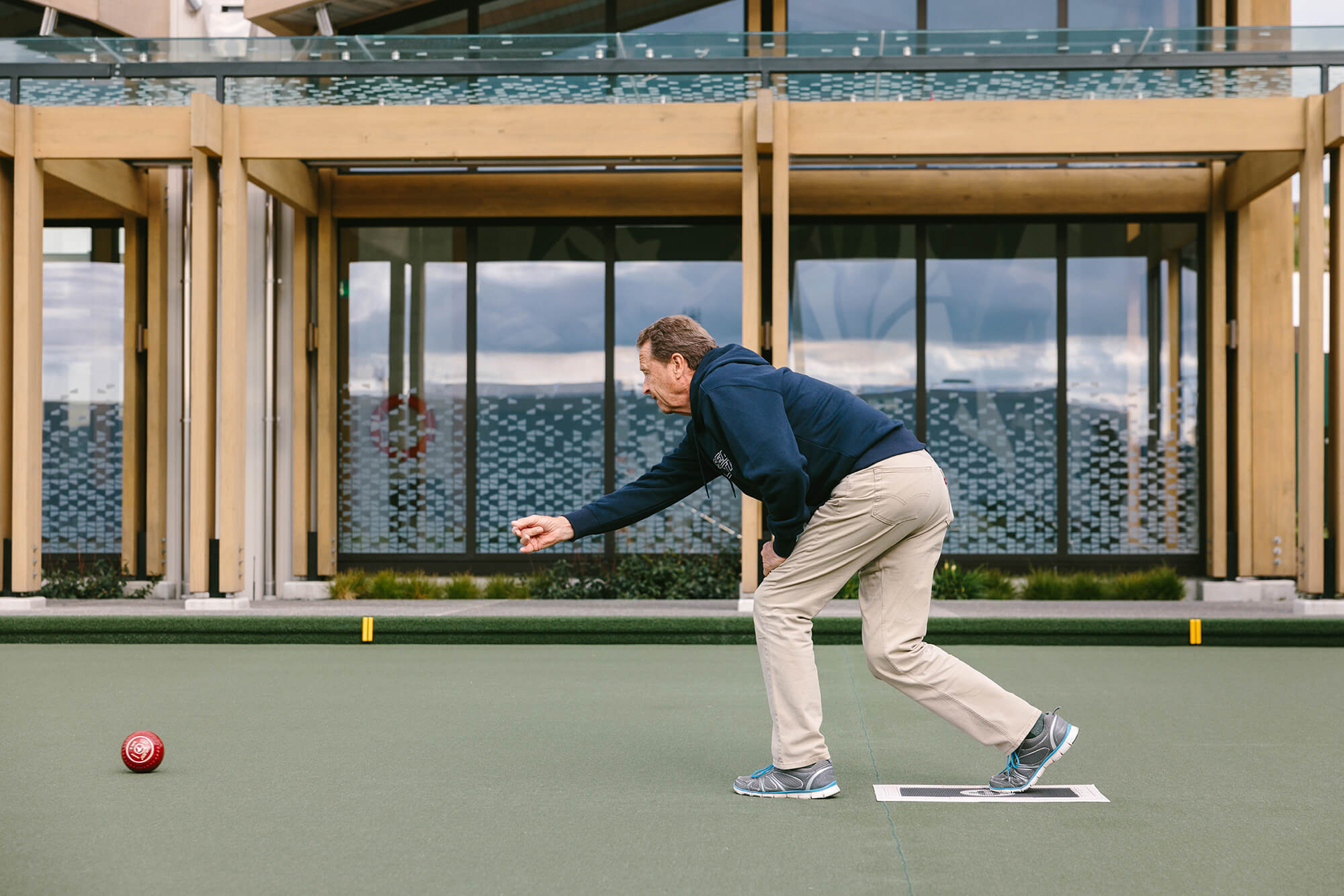 Man in a semi crouched position releases red bowl onto the rink.