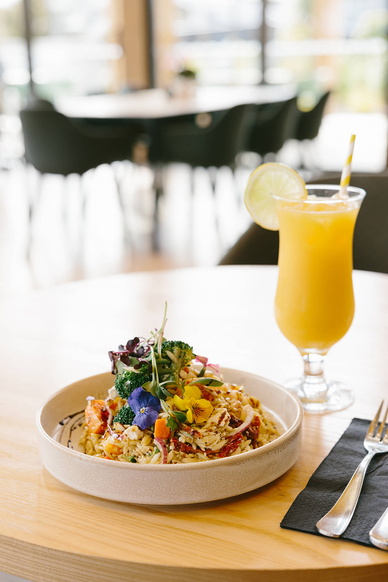 Bowl of orzo pasta salad topped with brightly coloured edible flowers. A cocktail glass of orange juice stands adjacent.