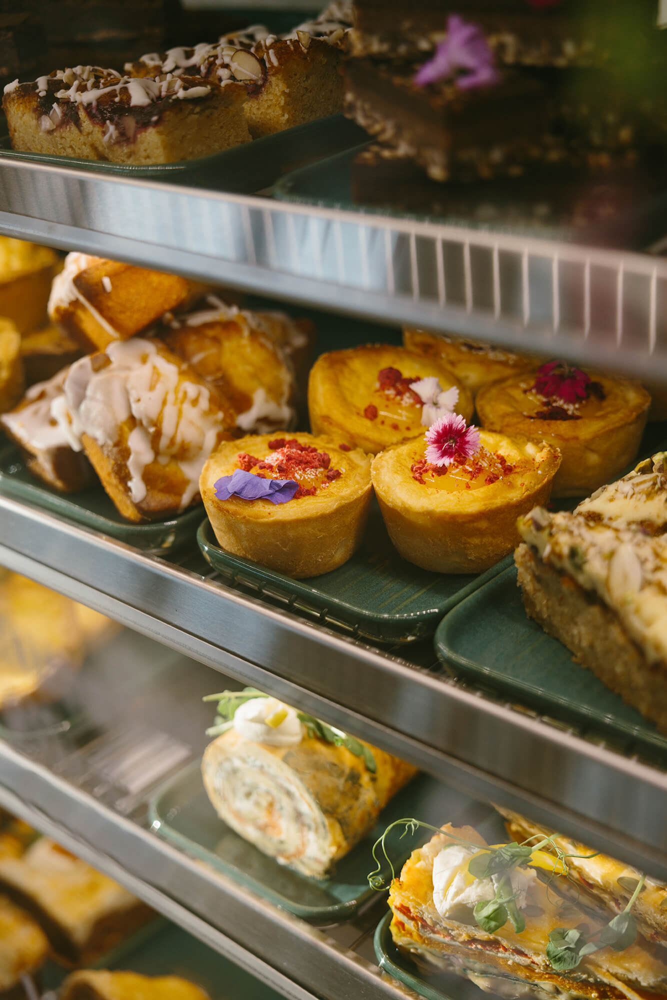 Cabinet food, including orange cakes, chocolate slices, lasagna and salmon rolls, viewed through cabinet glass.