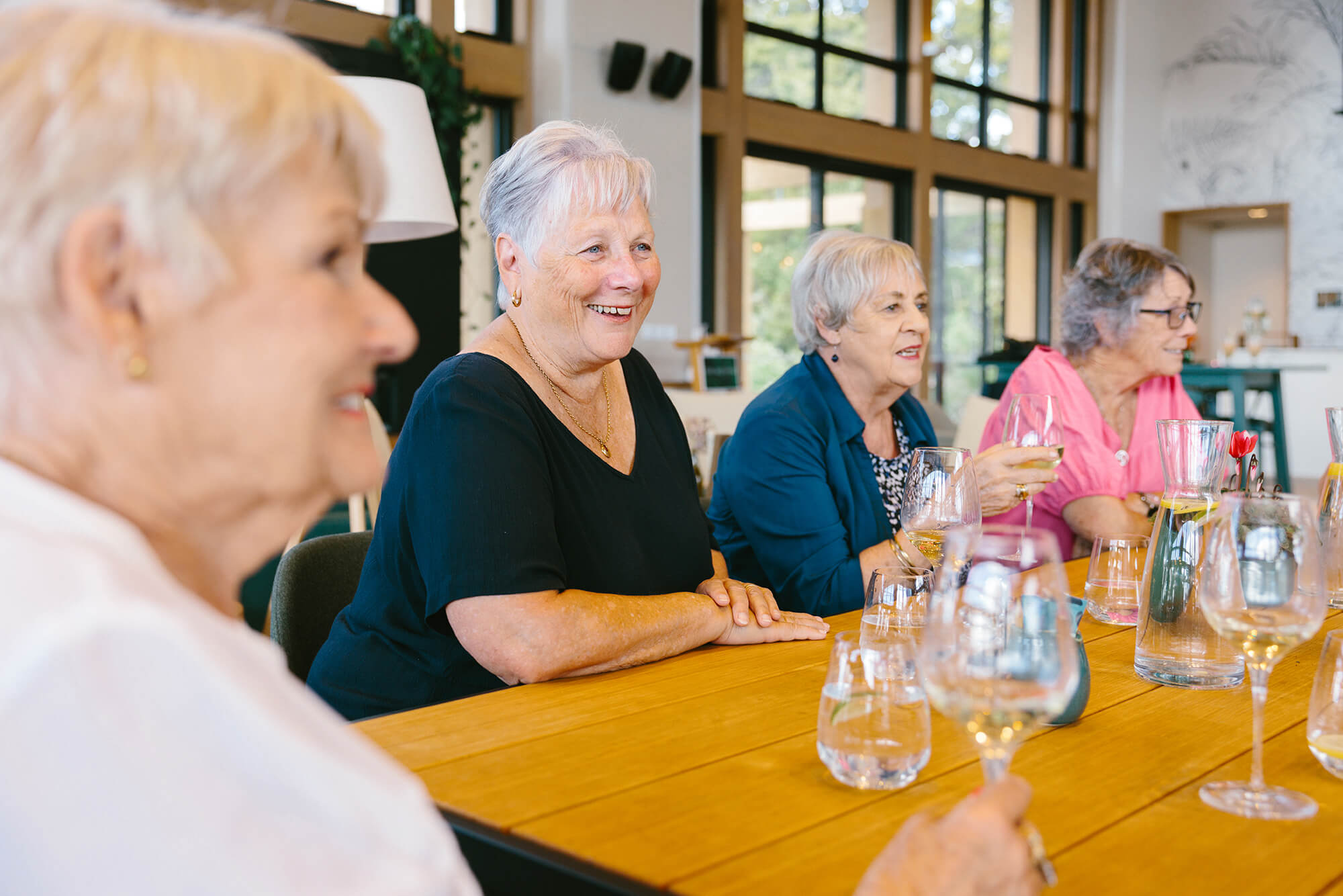 Smiling retired woman sit around a table holding glasses of wine, deep in conversation.