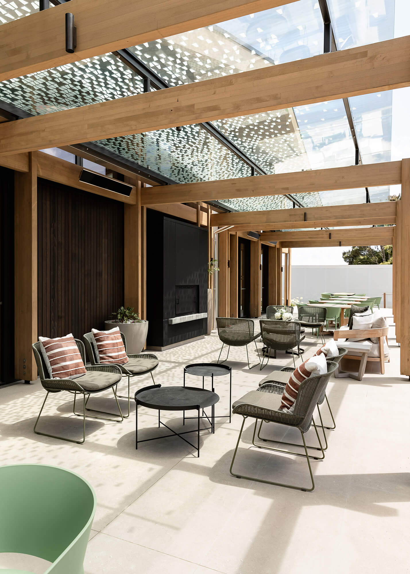 Cushioned outdoor seating and short coffee style tables on an expansive concrete area, beneath a pergola with glass roof.