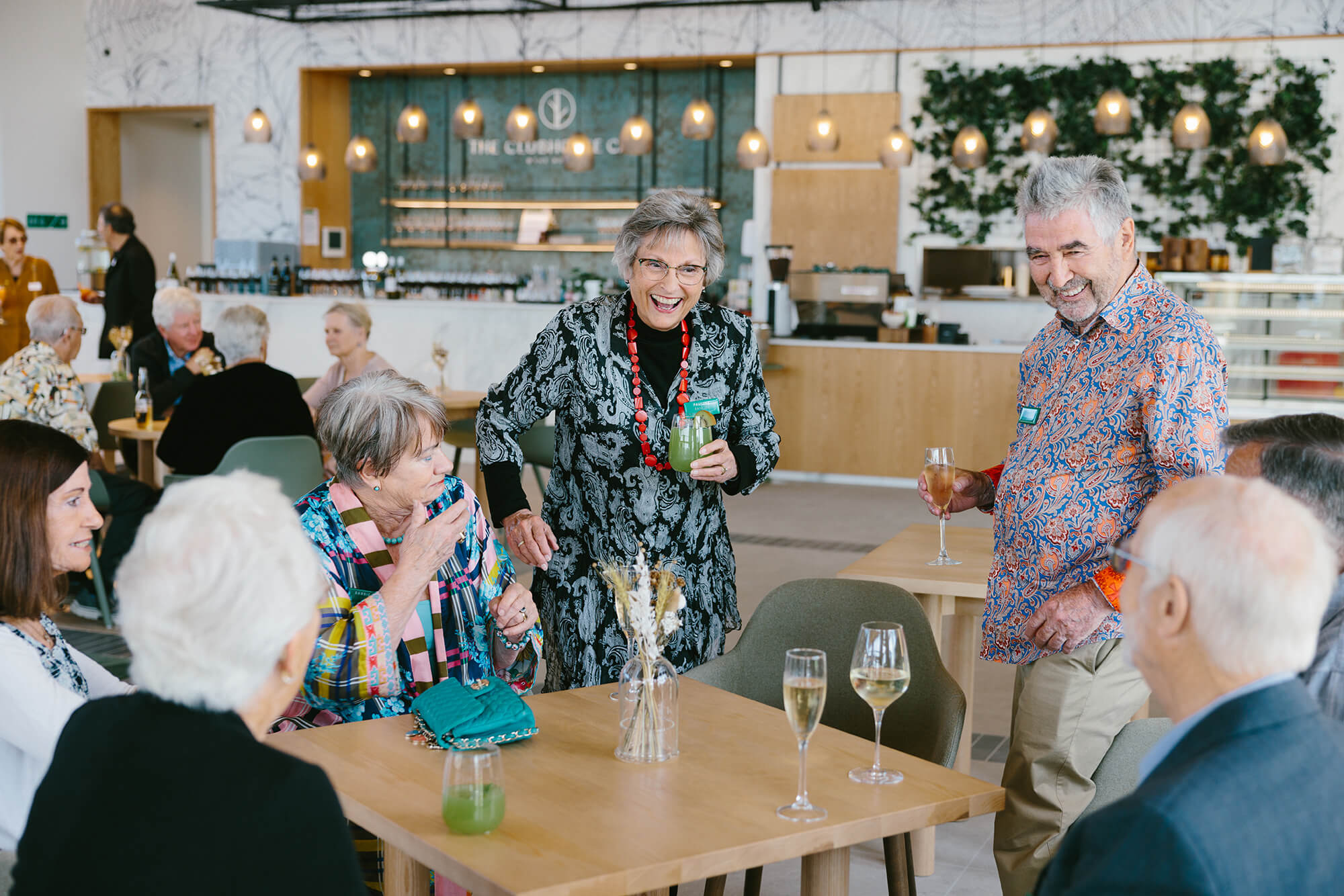 Group of retirees sit and stand around a wooden square table, holding alcoholic drinks and talking happily.