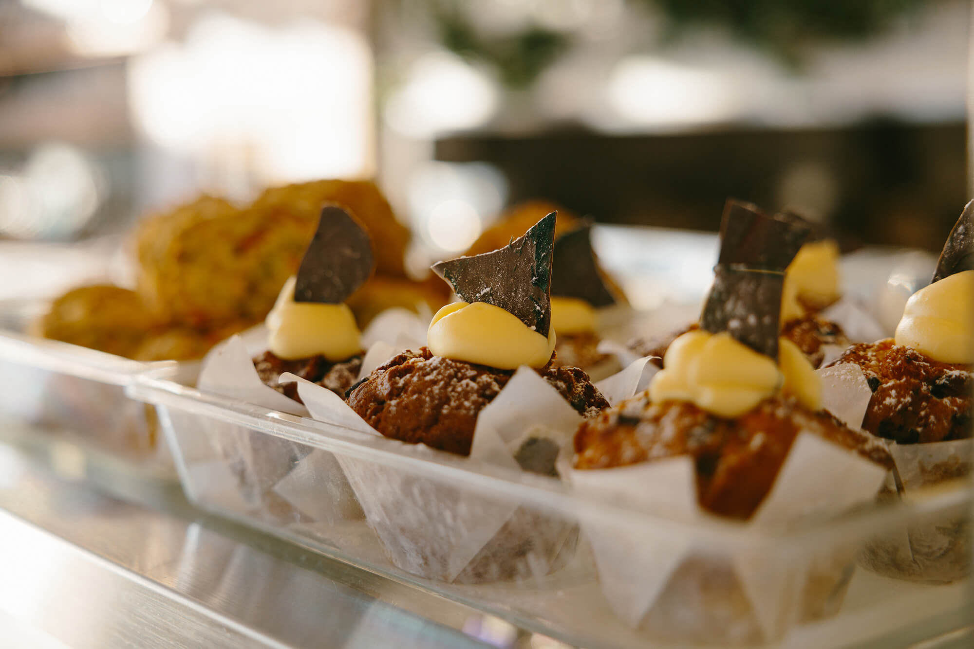 Glass tray of freshly baked muffins topped with yellow icing and a piece of chocolate.