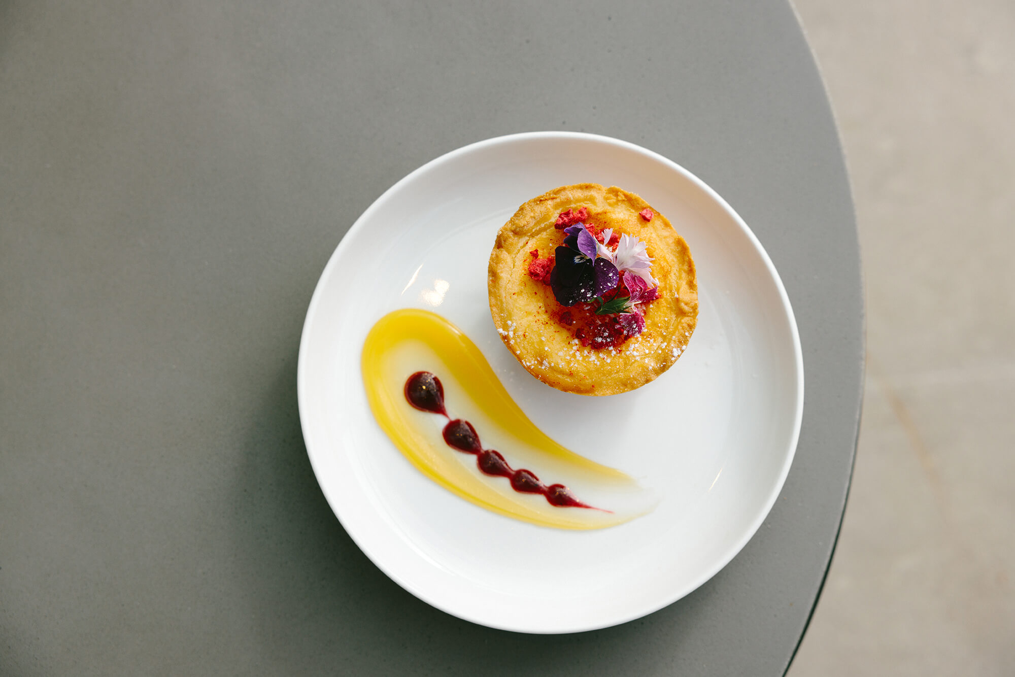 Birds eye view of an orange cake with purple edible flowers on top. Plated on a crisp white plate with a citrus compote swirled on the left.