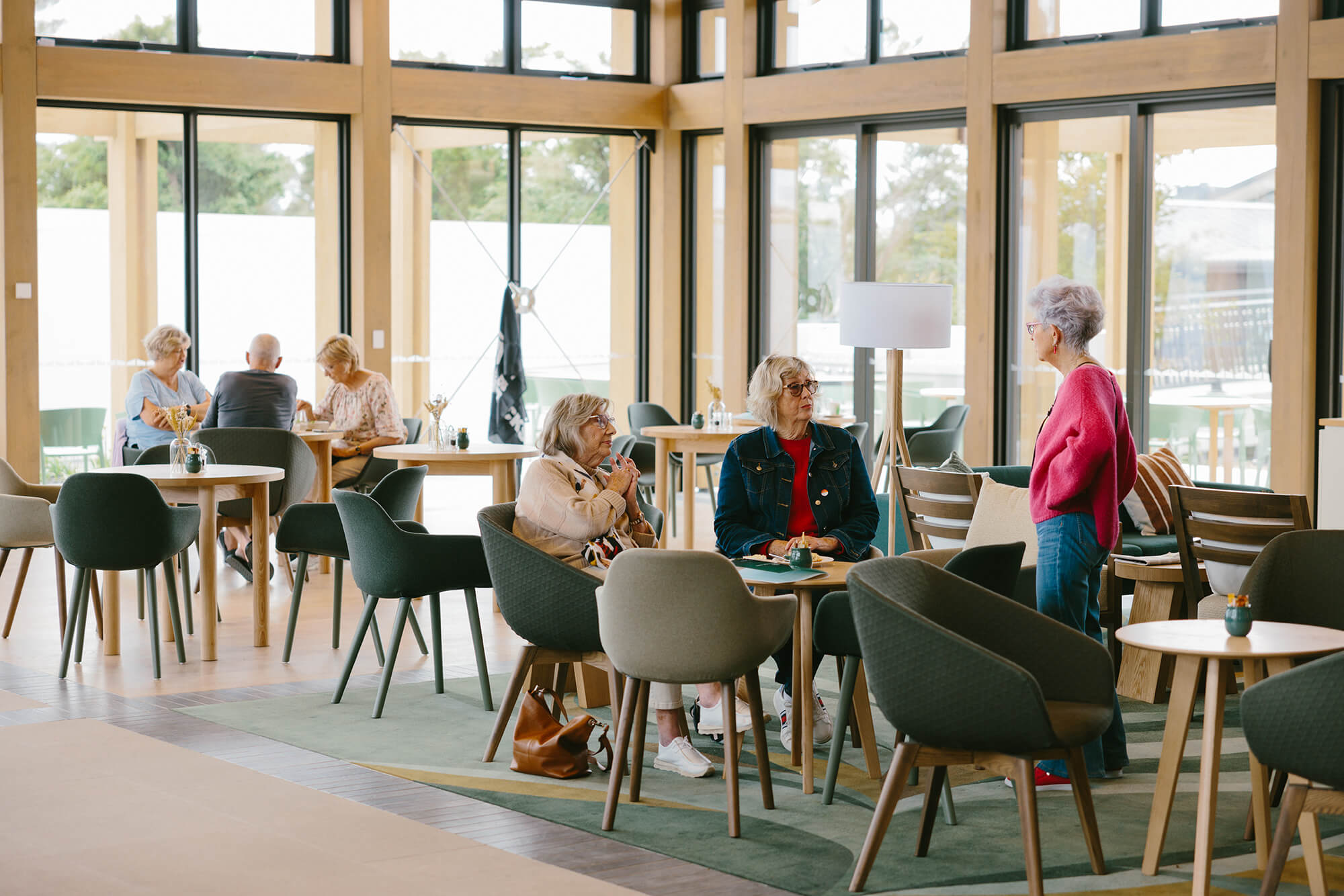 Two women sat at a clubhouse cafe table have a conversation with a woman standing close by.