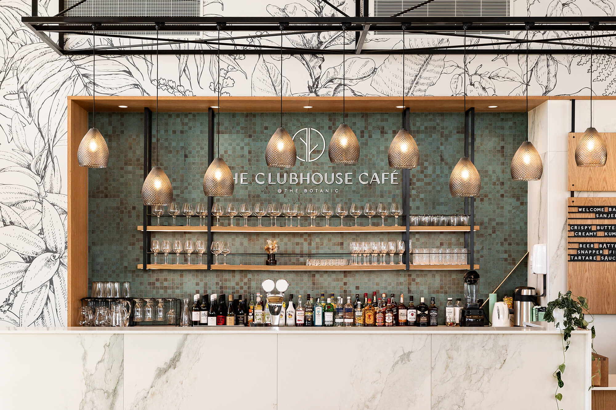 Pendant lights hang at varying heights above the serving bar. Glassware is organised on two shelves on the back wall, bottles of liquor sit below.