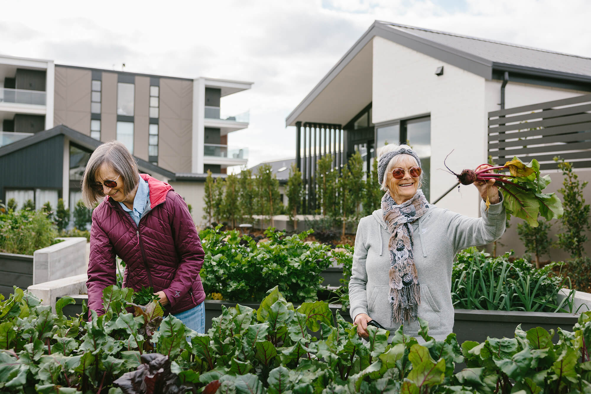 Two women stand by a raised vegetable garden, one woman holds up a freshly harvested beetroot.