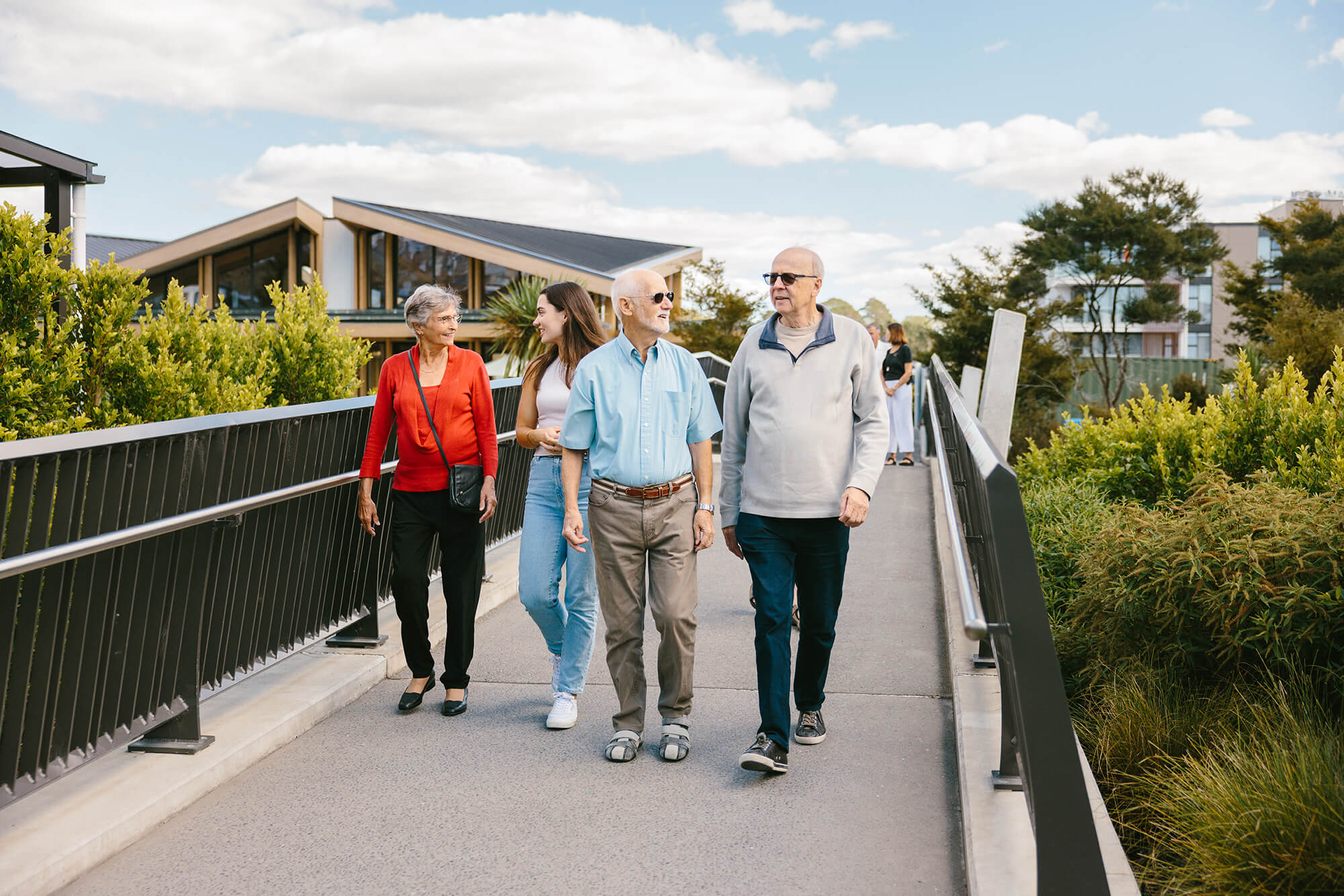 Multigenerational group of people visiting the village, walk across a bridge while in conversation with the clubhouse behind them.