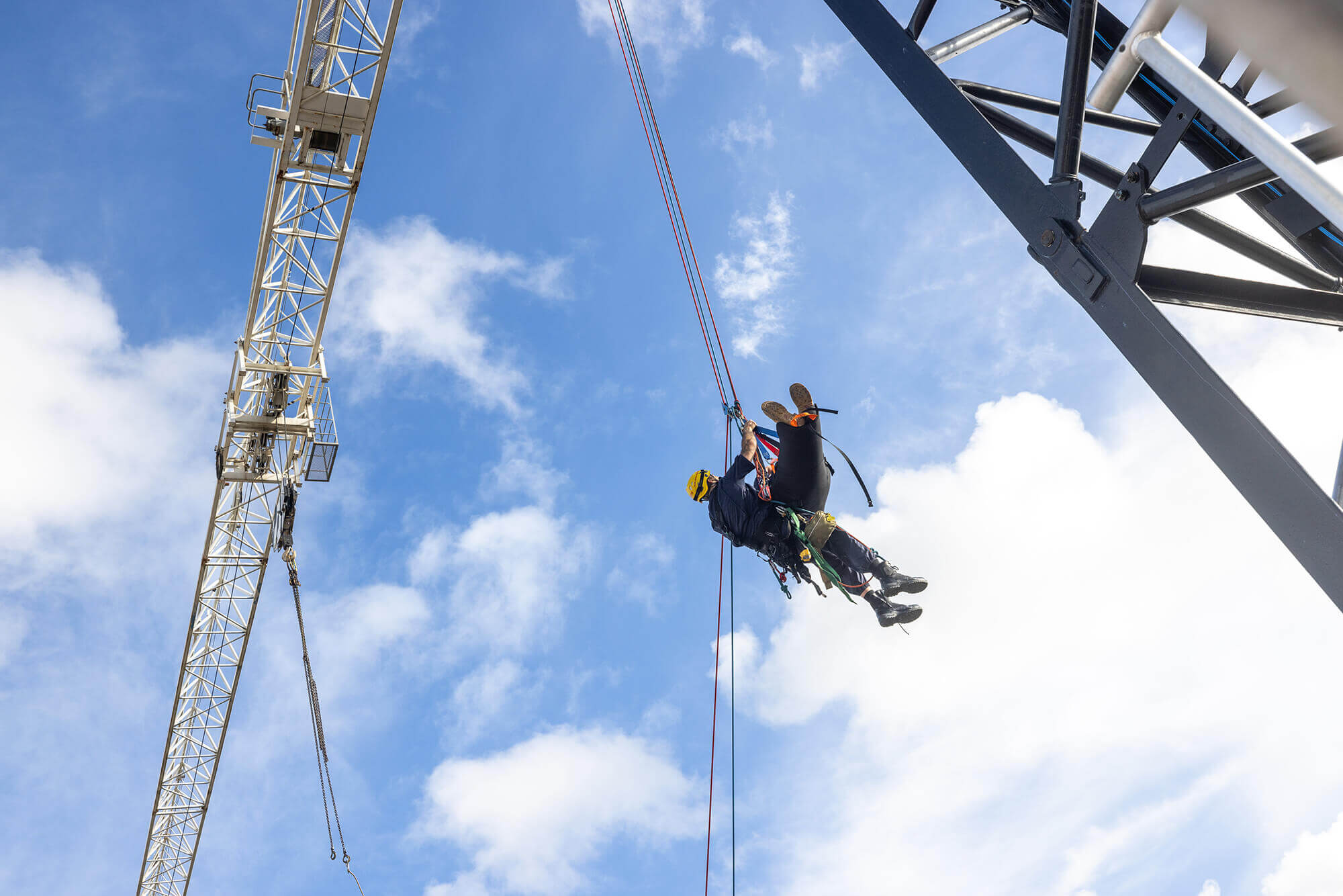 A fire brigade volunteer in a harness hangs in the air holding a recused crane operator in a safety sling. 