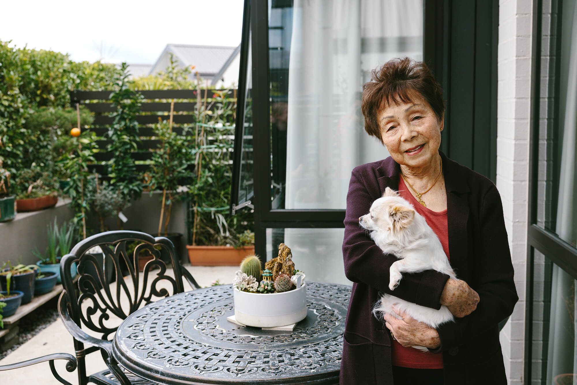 Retired woman stands in front of a black table on her patio holding a small white dog in her arms.