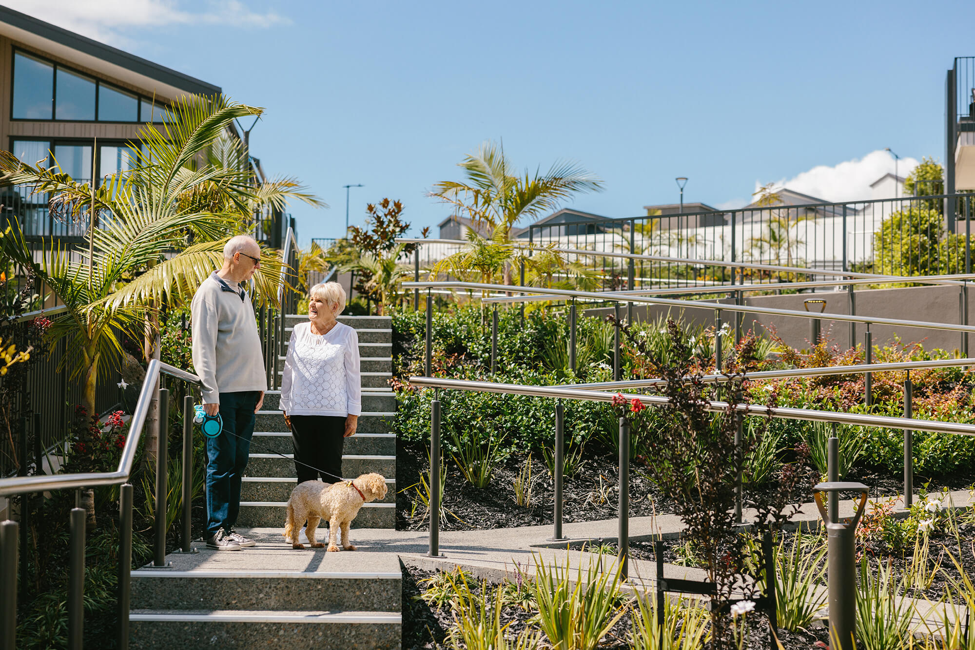 Two retired friends and a golden labradoodle stand on the landing between sets of outdoor stairs with lush plantings in the background.