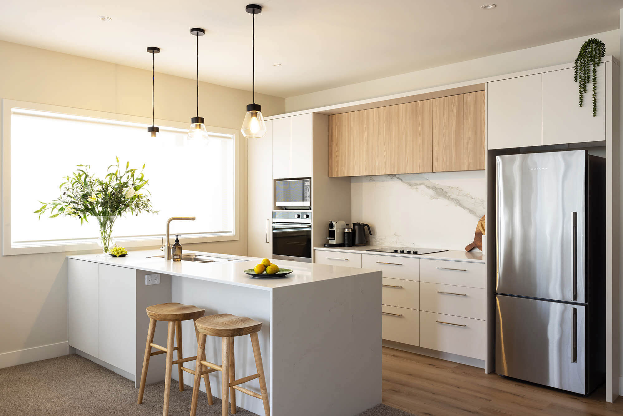 Sunny, open plan kitchen with light wooden cupboards and white drawers. Two bar stools sit under the island bench.