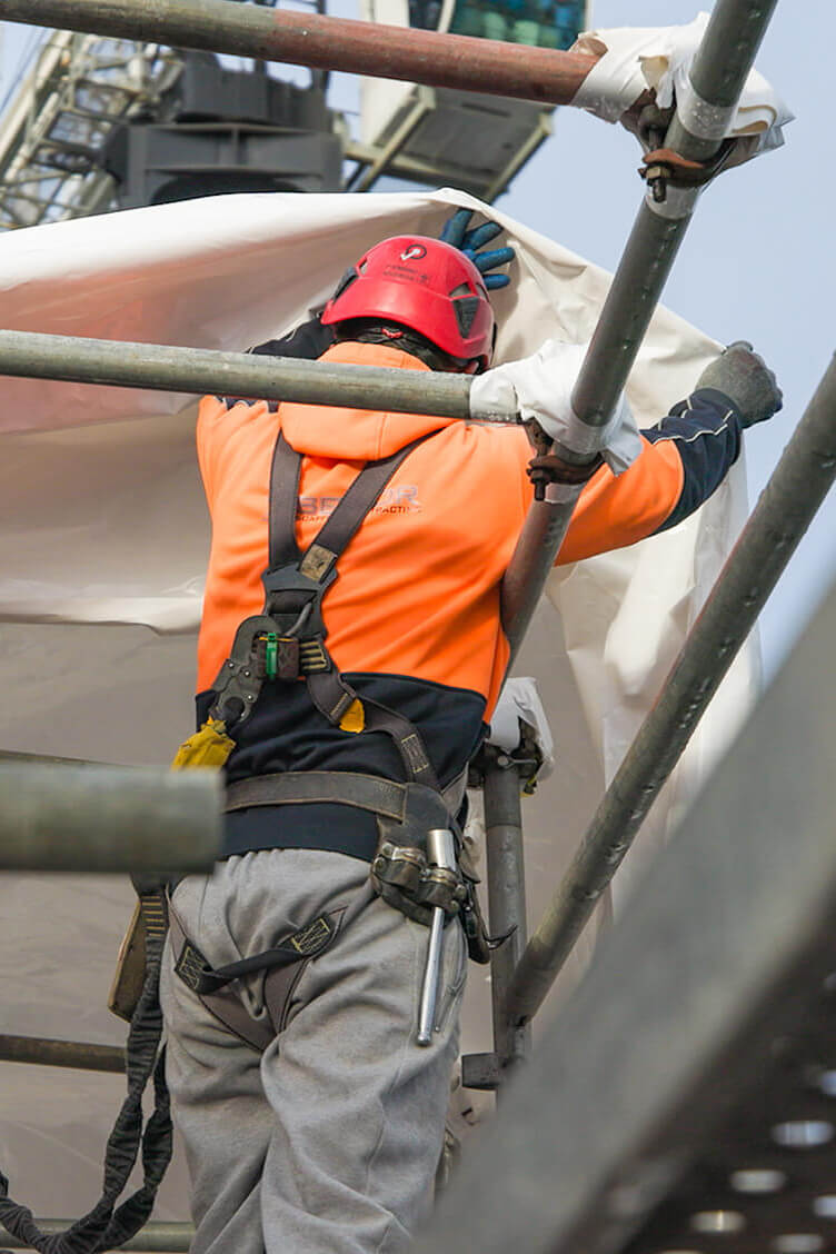 Man in PPE gear and safety harness pulls roof tent fabric over scaffold framing.