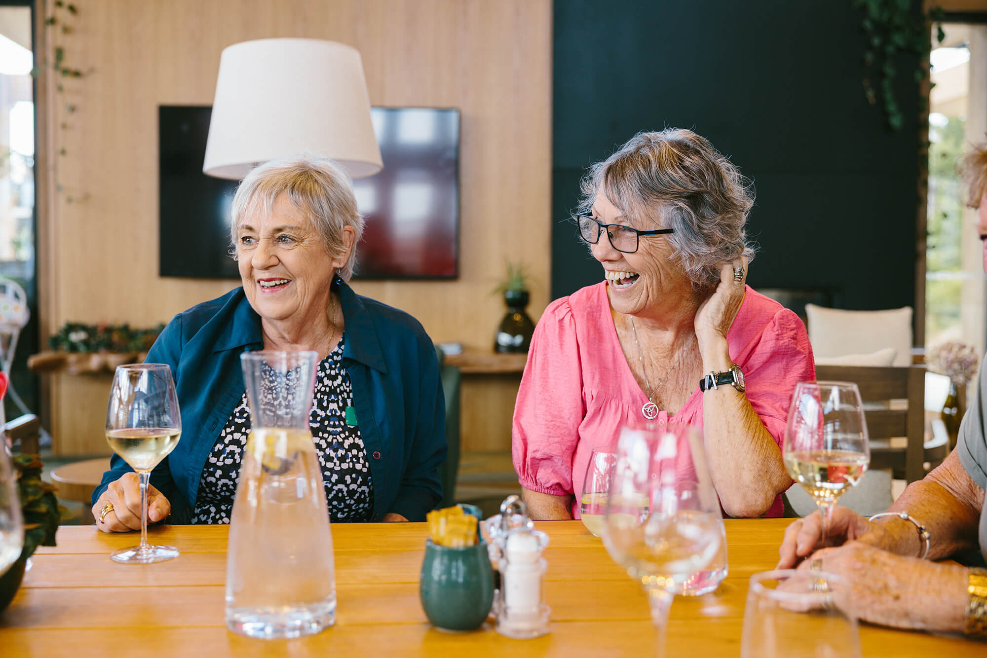 Two women sit at a table in The Clubhouse Café holding glasses of wine and laughing.