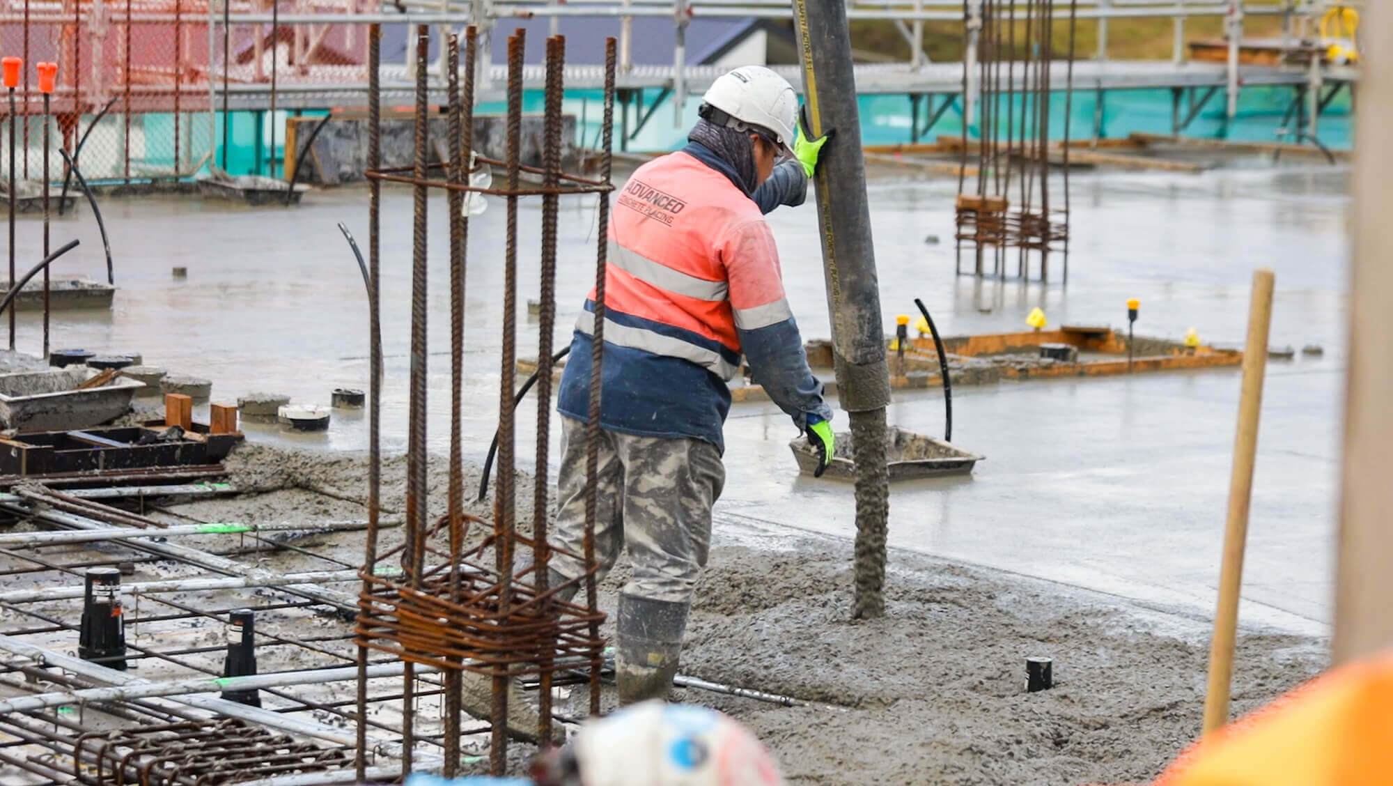 Man in PPE gear stands in a construction site while he directs tube delivering concrete into the slab.